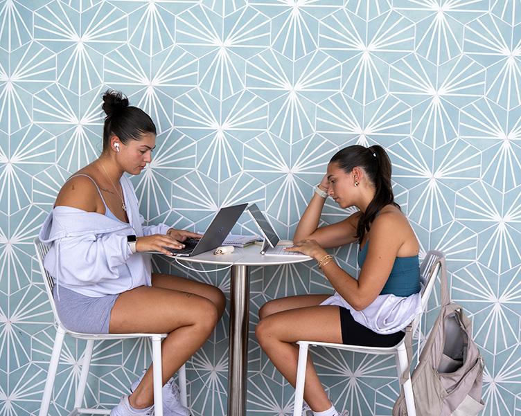 Two students working on laptops at a hightop table.